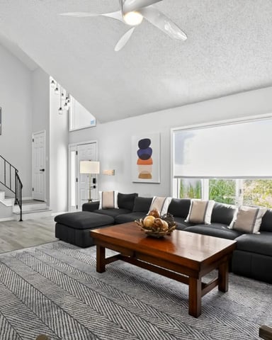 Bright living room with two blue sectional sofas facing a pool table, yellow brick fireplace with TV above, and large windows overlooking outdoor landscape.